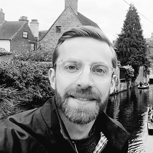 a black and white image of a man in glasses with a short beard in front of a canal. he looks happy.